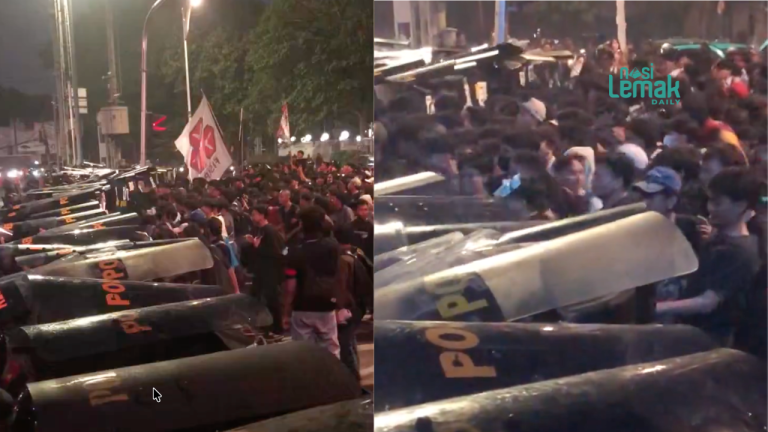 Indonesian riot police hold a barricade against a large crowd of protesters and students on an urban street at night.